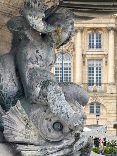 The image captures a close-up view of a weathered bronze sculpture of a cherub riding a fish, part of the Fontaine des Trois-Grâces (Fountain of the Three Graces) located in Place de la Bourse in Bordeaux, France. The sculpture exhibits significant patina and signs of age, with patches of verdigris and rust. The cherub is depicted in a dynamic pose, with one arm raised and the other resting on its leg. The fish sculpture beneath it is detailed with scales and a large fin. In the background, the classical architecture of the Place de la Bourse is visible, featuring ornate windows and balconies. Water is actively flowing from the fountain, creating a mist that is visible in the air. A few individuals are partially visible in the lower right portion of the frame, suggesting the fountain is in a public area.