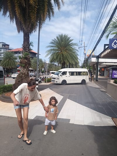 The image shows a woman and a young child walking on a crosswalk in Ban Ao Nang, Thailand. The woman is wearing a white t-shirt with a lobster graphic, denim shorts, and flip-flops. The child is wearing a brown t-shirt, light-colored shorts, and white clogs. They are holding hands. In the background, there is a white van, palm trees, buildings, and a street with motorbikes and cars. A McDonald's restaurant is visible further down the street. Overhead power lines are strung across the sky.
