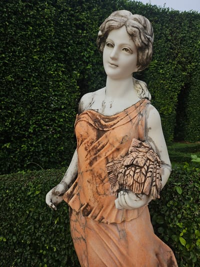A white marble statue of a woman in a draped orange garment stands in front of a dense green hedge wall in Na Chom Thian, Thailand. The statue depicts a young woman with her hair styled in curls, her head tilted slightly to the left, and her gaze directed upward. She is holding a bundle of what appears to be wheat or grain in her right arm. The statue shows signs of weathering, with dirt and discoloration visible on its surface. The hedge wall behind the statue is meticulously trimmed and forms a lush green backdrop. The ground in front of the hedge is also covered in low-lying green foliage. The sky above is overcast, suggesting a cloudy day. The overall scene is one of serene, classical beauty set within a cultivated garden environment.