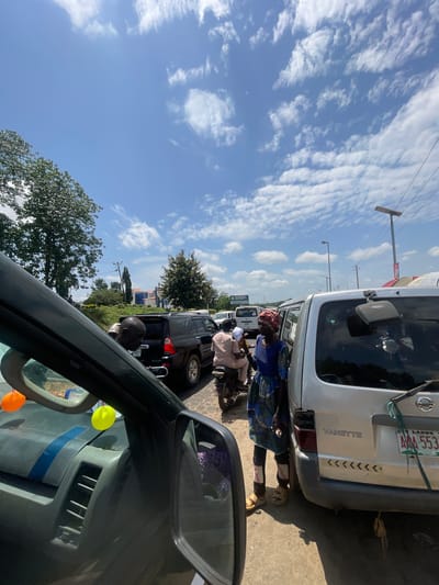 A street view captures a traffic scene on a sunny day. Several vehicles, including a black SUV and a silver van labeled "VANETTE," are stopped on a paved road. A person in a blue and green patterned dress and a red headscarf stands beside the silver van. Another person, wearing a light-colored shirt and pants, is seated on a motorcycle behind the black SUV. The background shows trees, buildings, and utility poles under a bright blue sky with scattered white clouds. A sign with the text "Gbongan University" is visible on the left side of the image. The perspective suggests the photograph is taken from inside another vehicle, with part of its dashboard and side mirror visible in the foreground.