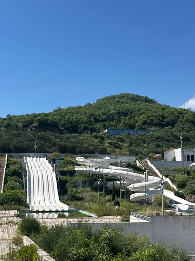 In Podostrog, Montenegro, a weathered amusement park stands against a backdrop of lush green hills. The park's centerpiece is a large, white water slide, its fluted surface descending into a murky pool. Surrounding it are several other tubular water slides, their white curves contrasting with the overgrown vegetation. Abandoned structures, some appearing to be part of a hotel or restaurant, are scattered amongst the slides, their windows dark and their surfaces stained. The sky above is a clear, vibrant blue. A faded sign on the hillside reads "AQUA PARK." The scene suggests a once-thriving attraction now reclaimed by nature.