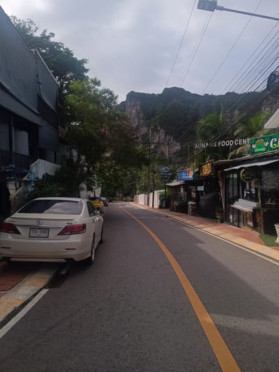 The image captures a street scene in Ban Ao Nang, Thailand. A cream-colored Toyota Camry is parked on the left side of the street, partially obstructing the sidewalk. The license plate is visible, reading "กม 1580 กระบี่". To the right, a row of shops and businesses lines the street, including what appears to be a food center called "AONANG FOOD CENT". Signs for "ROAD" and "CANNABIS" are also visible. Overhead, a dense network of power lines is strung across the sky. In the background, a large limestone cliff rises. The street has a yellow center line, and several other vehicles can be seen parked further down the road. The weather seems overcast.