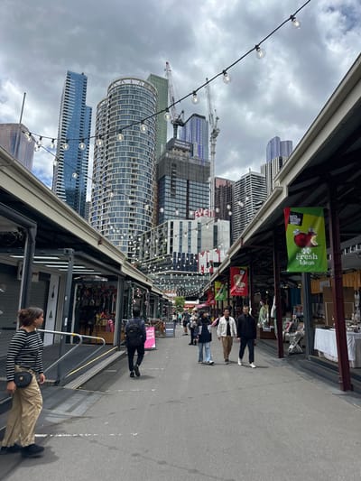 A bustling marketplace street in a modern city. On the right, pedestrians stroll along the walkway lined with shops. Above, banners advertising "Fresh Ideas" hang from a covered area. In the background, a view of modern skyscrapers under a cloudy sky is visible. String lights are strung across the street. Construction cranes dot the skyline amidst the tall buildings.