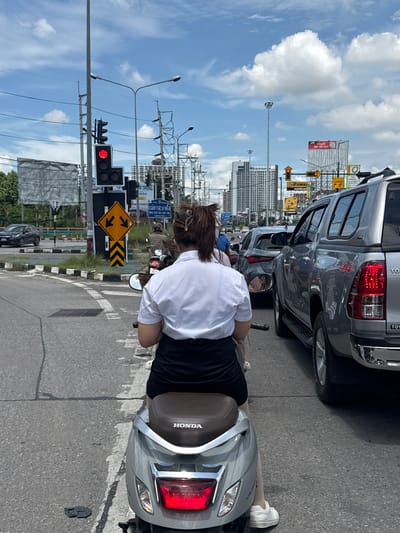 The image depicts a street scene in a city, possibly in Thailand given the presence of Thai text on signs. In the foreground, a person is riding a silver scooter, viewed from behind. The rider is wearing a white short-sleeved shirt and a black skirt. The scooter has a brown seat with the "HONDA" logo. In the middle ground, traffic is backed up, including a silver pickup truck to the right and other vehicles further ahead. Traffic signals are visible, with a red light illuminated. A yellow directional sign with arrows pointing left and right is also present. In the background, tall buildings and utility poles are visible against a sky with scattered clouds. The environment appears to be an urban intersection with multiple lanes of traffic. The location marker "บ้านกาญจน์กนก 23, Thailand" suggests a specific area within a city.