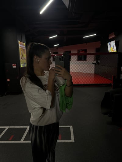 A woman stands in a dark gym in Vniissok, Russia, holding a smartphone. She is wearing a white t-shirt, dark pants, and has an earbud in her ear. A green bag is near her shoulder. The gym has a boxing ring with red accents, a television screen, and various workout equipment. Lights are visible on the ceiling. The floor is marked with white lines and red accents. A poster or sign is placed near the woman.