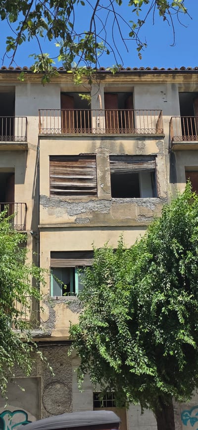 An exterior shot of a dilapidated building, likely abandoned, is captured in Huesca, Spain. The building features multiple stories with weathered facades and balconies. The balconies have rusty iron railings with a decorative criss-cross pattern. Some windows are boarded up with wooden slats, while others are open, revealing darkness within. One window on a lower floor has a light blue frame. Lush green trees with dense foliage partially obscure the lower sections of the building. In the foreground, the top of a dark grey car-top carrier is visible, with graffiti adorning the wall behind it. The sky above is clear and blue, with tree branches and leaves framing the top of the image.
