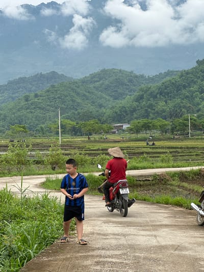 In Mai Chau, Vietnam, a young boy in a blue and black striped soccer jersey and black shorts stands on a dirt path. To his right, a person wearing a red shirt and a conical hat rides a red motorcycle. The path leads towards a verdant landscape of rice paddies with terraced fields, where several people are working. In the background, rolling green hills rise up, partially obscured by a cloudy sky.