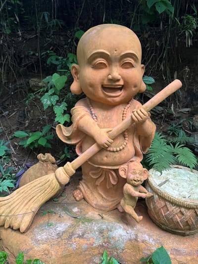 A full shot shows a terracotta statue of a laughing child monk holding a broom in a courtyard. The monk statue is positioned in the center of the frame, kneeling on a large rock and smiling broadly with his eyes closed. He wears a beaded necklace and is holding a long, wooden broom with his arms crossed. To the left of the monk, a small, brown sack with a rope tie rests on the rock. To the right of the monk, a small, mischievous-looking monkey statue stands with its hands on a woven basket. The background consists of dense green foliage and dark, rocky terrain. The overall scene is vibrant and playful, suggesting a garden or outdoor decorative setting in the Tây Hoa Lư Ward of Vietnam.