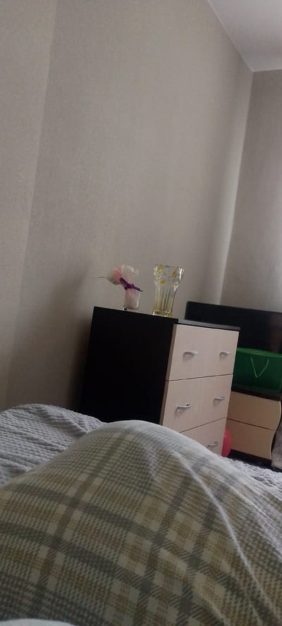 This is a slightly blurry, eye-level, indoor, medium shot of a bedroom scene. In the foreground, a person is under a grey and white plaid blanket, with only the blanket visible. The blanket is bunched up, suggesting the person is lying down. Behind the blanket, to the right, is a light wood dresser with three drawers, each with a silver handle. The dresser has a black top. On the black top of the dresser, to the left, is a small glass vase with a light pink flower with a purple ribbon. To the right of the flower is a clear glass vase with a yellow floral pattern. To the right of the dresser, there is a green tote bag with a white "V" shape on it. The background consists of a textured, light grey wall that curves upwards, suggesting a corner of the room or a ceiling. The lighting is soft and diffused, creating a gentle shadow on the wall behind the dresser. The overall scene appears to be a relaxed, domestic setting. The location is a bedroom in Volzhsky, Russia.