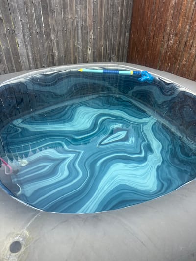 The image displays a close-up, overhead view of a surfboard with a mesmerizing, abstract blue and white marbled design, reminiscent of agate. The surfboard rests on a wooden deck, with planks of varying shades of brown and grey visible to the left. A blue cleaning tool, possibly a pool skimmer or brush, is attached to the side of the surfboard, its bright yellow tip and blue handle adding a pop of color against the patterned surface. The scene appears to be outdoors, under natural light, suggesting it is daytime. The reflection on the surfboard indicates a smooth, possibly wet surface. The location cues, such as the wooden deck, suggest an outdoor setting near water or a pool area. The overall impression is one of leisure and outdoor activity.