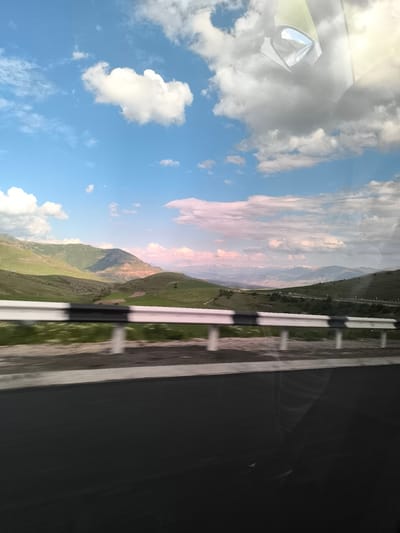 The image captures a panoramic view of the mountainous landscape of Yelpin, Armenia, taken from a moving vehicle. The scene is dominated by a vast blue sky, dotted with fluffy white cumulus clouds and larger grey cloud formations. The light appears soft, possibly late afternoon, with subtle pink and golden hues on the distant clouds and mountains.
In the mid-ground, rolling green hills and mountains stretch across the horizon, some displaying darker, rugged patches. Further in the distance, snow-capped peaks are faintly visible, adding to the grandeur of the natural setting.
The foreground features a black asphalt road, suggesting a highway or a well-maintained route. A white and black striped guardrail runs along the side of the road, exhibiting motion blur, which indicates the photograph was taken while traveling at speed.
A notable detail is the clear reflection in the upper right quadrant of the image. It shows parts of a vehicle's interior, specifically a side mirror and what appears to be elements of the car's ceiling or windshield, confirming the perspective from inside a moving car. This reflection suggests the photograph was taken through a window.
The overall impression is one of a scenic journey through a picturesque, mountainous region under partly cloudy skies. There is no visible text in the image.