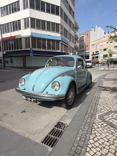 A light blue Volkswagen Beetle is parked on a street in Aveiro, Portugal. The car's license plate reads "DN-18-97". The location appears to be a commercial area, with tall buildings, shops, and cobblestone sidewalks. A building with numerous windows is visible on the left side of the car. A street sign with a left-pointing arrow is mounted on the building. There is a storm drain next to the car. Other buildings are in the background. The weather seems sunny.