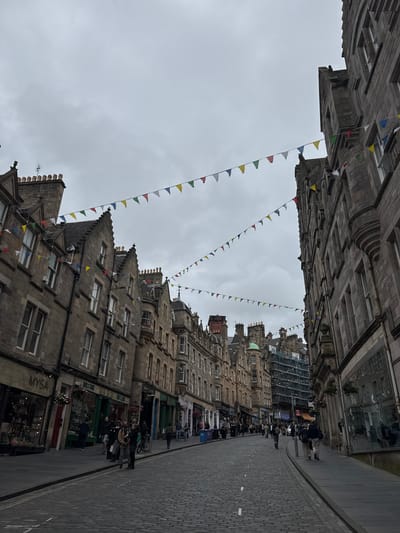 A street scene in Edinburgh, United Kingdom, features a cobblestone road lined with multi-story stone buildings. Strung between the buildings are triangular flags in various colors, including red, yellow, green, and blue. The sky is overcast and gray. Several people are visible walking along the sidewalks and crossing the street. The buildings have traditional stone facades with numerous windows and decorative architectural elements. Some of the ground-floor businesses have awnings and shop windows displaying goods. Scaffolding is present on one of the buildings further down the street, indicating ongoing construction or renovation.