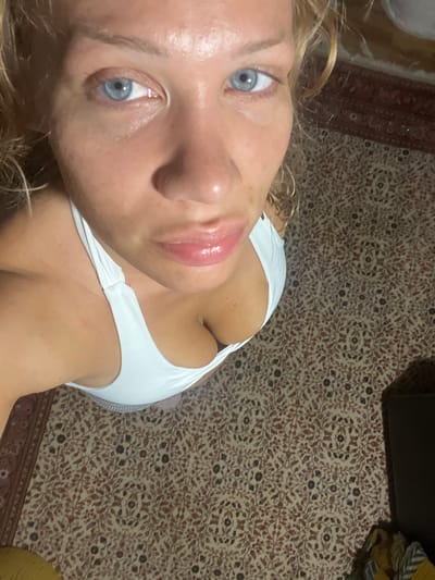 This is a close-up, overhead shot of a young woman with blonde hair and blue eyes. She is wearing a white top. Her face is tilted upwards towards the camera, and her lips are parted slightly, with a glossy appearance. The background is a patterned carpet with a reddish-brown and cream color scheme. The lighting suggests an indoor setting, possibly with artificial light as there are no strong shadows indicative of natural daylight. The overall mood is casual and intimate due to the close-up nature of the shot and the direct eye contact. There is no visible text in the image. Given the location context of Mühldorf a. Inn, Germany, the setting is likely a domestic interior within that region. The image does not provide information about the time of day or weather conditions.