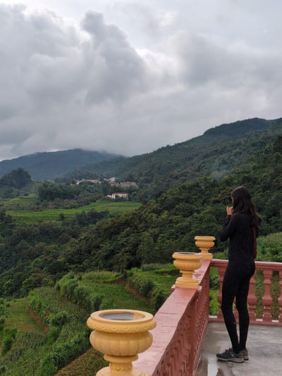 A person stands on a balcony overlooking a mountainous, verdant landscape in Lũng Cú Commune, Vietnam. The sky is overcast and cloudy, with the clouds appearing heavy and gray. The person, facing away from the camera, wears black athletic clothing and dark sneakers. They appear to be holding a device to their face, possibly a camera or phone, and are looking out at the scenery. The balcony features decorative red railings and large, ornate yellow planters. In the background, lush green hills and mountains dominate the view. Terraced fields, characteristic of the region, are visible on the slopes, and scattered buildings suggest a small settlement nestled within the landscape. The ground on the balcony appears wet, indicating recent rain.