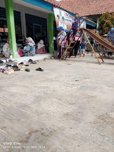 A group of people gathered outside a building with blue-trimmed windows and a red-tiled roof. Some sit on the building's porch while others stand near a playground slide. A child is on the slide. Shoes are scattered on the ground near the building. A banner hangs from the building. A woman wearing a hijab is near the slide. The ground is concrete and shows signs of wear.