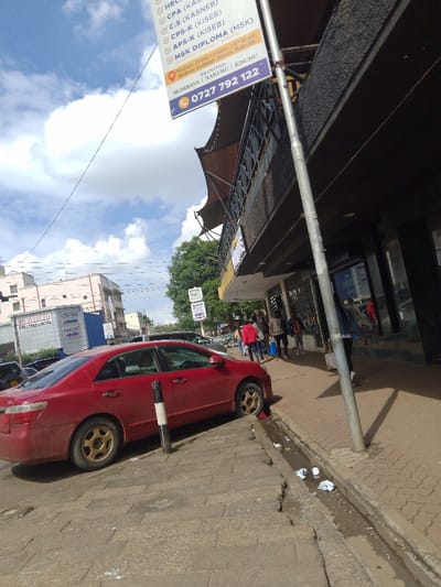 The image shows a red sedan parked on the street in front of a shop. The shop is on the right side of the image, and a street sign is visible above the car. The sign advertises a school, MSK Diploma, with phone numbers and branch locations. The street has a sidewalk and a gutter running along its edge. There are people walking on the sidewalk. The sky is blue with white clouds. The scene is located in Athi River, Kenya, at the coordinates -1.453 latitude and 37.007 longitude. The car is facing away from the camera, and the front right wheel is turned slightly to the left. The car's rear tire is visible. The sidewalk is made of gray concrete pavers. There are some pieces of trash in the gutter. The weather appears to be sunny.