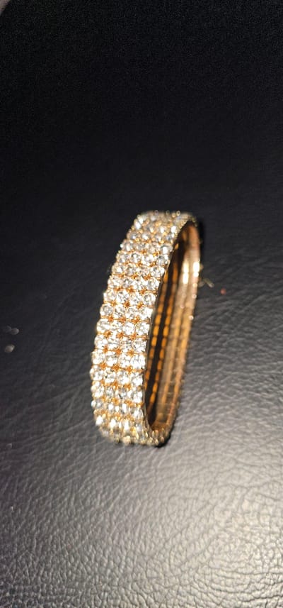 A close-up, high-angle shot showcases a sparkling bracelet with three rows of clear, faceted stones set in a gold-toned metal band. The bracelet is positioned on a dark, textured surface that appears to be faux leather, with a visible grain pattern. The lighting creates bright reflections on the stones and the metal, suggesting a well-lit environment, possibly indoors. The bracelet is the sole object in focus, with the background slightly blurred, drawing attention to its intricate details and glittering appearance. No people are visible in the frame. The image does not provide any temporal or atmospheric cues like time of day or weather. There is no visible text in the image. The overall impression is one of luxury and ornamentation. The location context of Makati, Philippines does not offer specific visual cues within the image itself, but suggests an urban setting where such an item might be found or displayed.