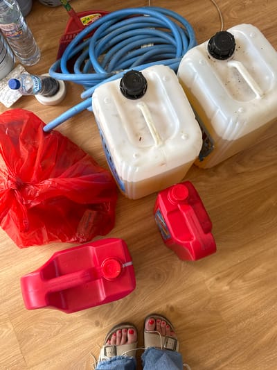 The image depicts a wooden floor littered with various items. Two white plastic jerrycans with black caps are stacked on top of each other. A coiled blue hose and a red broom head are nearby. Several plastic water bottles are visible in the lower left corner. Two red plastic gas cans and a tied red plastic bag are also present. A person wearing sandals and denim jeans is standing on the floor to the right. The scene is located in Nusa Dua, Indonesia.