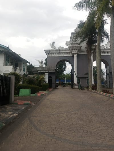 An outdoor scene in Suddepalle, India, captured under an overcast sky, depicts the ornate entrance to a property. In the foreground, a patterned paved driveway, resembling cobblestones, extends towards a large, multi-arched gate structure. The driveway's edges are bordered by curbs painted with alternating green and reddish-brown segments. To the immediate left, a black metal gate and a landscaped area with manicured green hedges are visible.
The central feature is a grand entrance gate structure, constructed from dark grey material with prominent white classical columns and decorative moldings. Multiple arches are integrated into the design, with a primary central arch serving as the main entry point. Elevated on the gate's roofline are several white sculptural figures, including at least two distinct winged creatures resembling eagles or angels. Tall palm trees are positioned to the right of the gate, partially obscuring the structure.
To the left of the gate, a multi-story white building with a dark, sloped roof and multiple windows and balconies is partially visible. Green foliage and various trees are present behind and around the gate structure. In the background, beyond the gate, a blue fence or wall is discernible. One individual on a motorcycle is observed in the distance, moving through one of the arches of the gate structure.