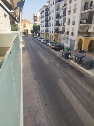 The image shows a street scene in Jerez, Spain, likely taken from a balcony or elevated position. A multi-lane asphalt road runs through the center of the frame, with parked cars lining the right side. Several residential buildings with white facades and balconies are visible on the right, with arched yellow storefronts at street level. A green tree is situated between two buildings. On the left side of the image, a modern building with a light-colored facade and glass railings is partially visible, suggesting a contemporary structure adjacent to older architecture. In the distance, a traditional building with an orange tiled roof stands out. Three large, dark gray trash receptacles are present on the sidewalk near the parked cars. A person in a blue top and dark pants is walking near the storefronts, and a silver scooter is parked to the right of the trash receptacles. The scene is illuminated by bright daylight, casting shadows across the road.
