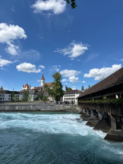 This is a photograph of the Aare river in Thun, Switzerland. The river flows rapidly in the foreground, with white water indicating strong currents. To the right, a historic wooden covered bridge spans the river, its weathered planks and roof visible. Flower boxes with red and green foliage adorn the sides of the bridge. In the background, the city of Thun rises, dominated by a large medieval castle with prominent towers and red-tiled roofs. A church with a tall, white steeple and a clock face is also visible. Buildings in the city appear to be a mix of modern and older architecture. A stone embankment runs along the riverbank, with a walkway on top where several people are casually strolling or standing. The sky is a vibrant blue, dotted with fluffy white clouds, suggesting a clear and sunny day. A tree branch with green leaves hangs into the frame from the top left corner.