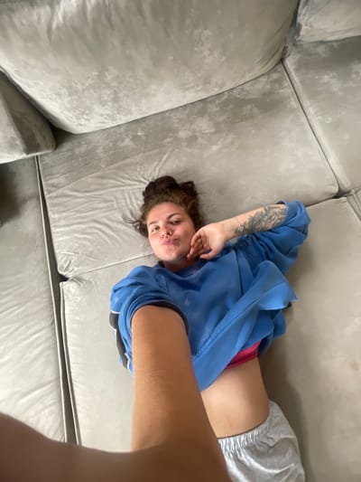 A woman with dark, messy hair pulled up in a bun is lying on her back on a light gray, plush sofa. She is wearing a bright blue sweatshirt, a pink bra, and light gray patterned shorts. A tattoo is visible on her left arm. Her right arm is extended towards the camera, as if taking a selfie. The woman is making a puckered face towards the camera. The sofa appears to be in a domestic setting, with no discernible external environmental features.