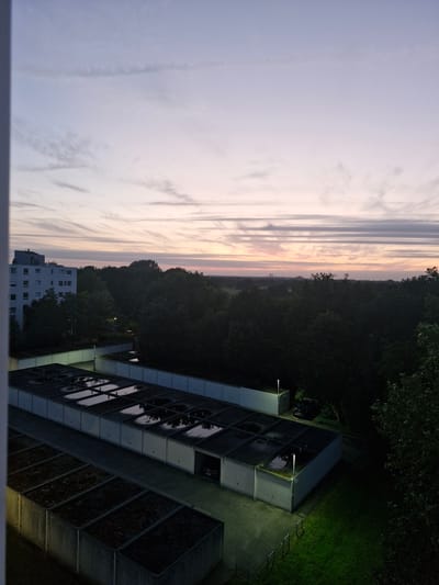 The image shows a twilight view of a residential area in Bremen, Germany. In the foreground, there are rows of concrete garage structures. The roofs of some of these garages have puddles of water, reflecting the sky. Beyond the garages, a dense line of dark green trees forms a natural boundary. To the left of the trees, a multi-story apartment building with white walls and numerous windows is visible. In the distance, the sky is a gradient of pale blue and pink, with wispy clouds streaking horizontally. The overall impression is one of a quiet, residential evening in a suburban or semi-urban setting.