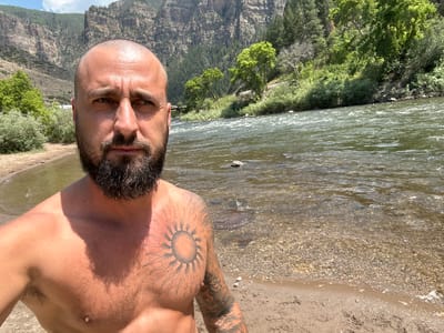 A bald, bearded man is taking a selfie on a sandy riverbank. He is shirtless and has a tattoo of a sun on his chest and other tattoos on his arm. The river is wide and flowing, with a rocky bed visible through the clear water. Lush green trees and steep, rocky cliffs line the opposite bank of the river. The sky is clear and blue with scattered white clouds. The image appears to have been taken on a sunny day. The location is identified as No Name, United States.