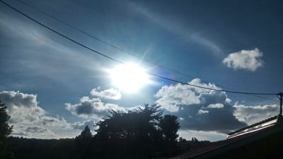 The image displays an outdoor scene in Ndeiya, Kenya, captured during daylight hours. A bright, central sunburst with associated lens flare dominates the upper-middle sky, which is predominantly blue with scattered white and grey cumulus clouds. Two dark, horizontal utility wires span the width of the frame in the upper section. The foreground features the upper portion of a reddish-brown corrugated metal roof on the right, connected to a dark utility pole from which wires extend. Below the wires, the middle and left foreground are occupied by dark, silhouetted trees and dense foliage. A distant, rolling landscape covered in trees is visible along the horizon line beneath the clouds. No people or overt actions are depicted.