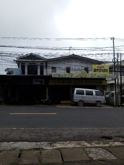 The image depicts a street-level view of a commercial building and roadway under an overcast sky. The user identified the location as the city of Neerveli, Sri Lanka; however, prominent signage on the building reads "BAKSO SOPONYONO," indicating an Indonesian business.
The scene features a two-story building in the midground, with a dark, recessed ground floor and a lighter, residential-style upper floor. Two large, rectangular yellow signs with green lettering and a central green circular logo are affixed to the facade, primarily displaying "BAKSO SOPONYONO." The ground floor consists of two dark storefront openings. A silver compact van with five windows per side is parked facing left on the shoulder in front of the right storefront. Two motorcycles are visible parked inside the dark recess of the left storefront. Overhead, numerous electrical and communication lines crisscross the frame against the bright, overcast sky. To the far right, a utility pole with additional lines and a section of another building are visible. In the foreground, an asphalt road with a single yellow center line extends horizontally, bordered by a concrete sidewalk composed of square pavers with weeds growing in the joints. No individuals are distinctly visible.