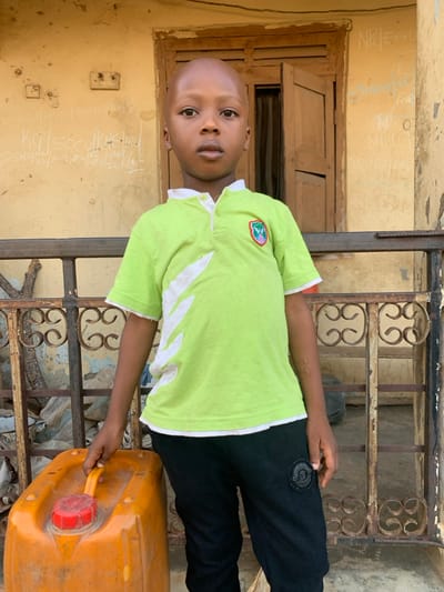 An image captured in Okene, Nigeria, depicts a young, dark-skinned child standing outdoors in front of a weathered building. The child, appearing to be male with a shaved head, faces the viewer directly with a neutral expression. He is wearing a light green short-sleeved polo shirt with white collar trim and white diagonal patterns on the sides, featuring a colorful emblem on the left chest. He also wears dark trousers, with a circular patch visible on the right thigh. To the child's left, an orange plastic jerrycan with a red screw cap and yellow handle rests on the ground, with the child's left hand resting on its handle.
The background consists of a light brown building wall showing discoloration and wear. A wooden window frame with two open shutters is visible behind the child. A dark, possibly metal, railing with decorative scrollwork elements separates the child from the wall. Various white inscriptions and scribbles are present on the wall, including "K/G | SÔC J.K.H.R.D" to the left and "N.P.EA.DE," "Shana," "H OT," and "NiGo" to the right of the child. The scene is illuminated by natural light.