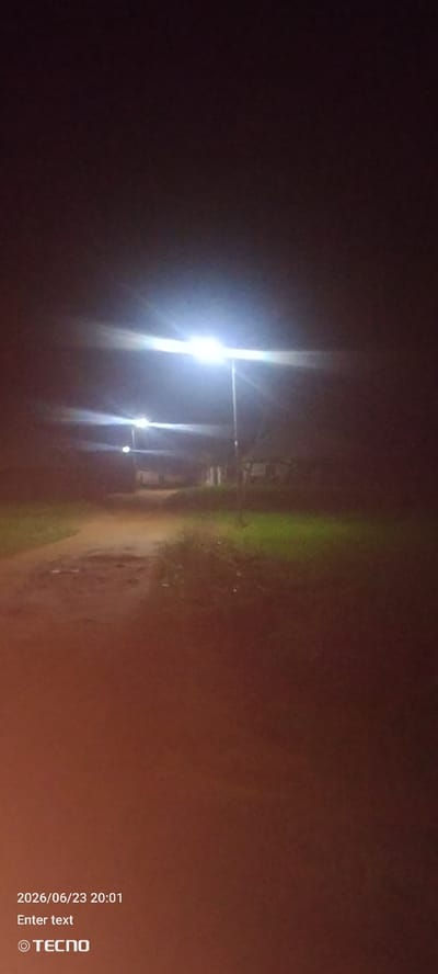 The image is a nighttime photograph of a dirt road illuminated by streetlights in Gumel, Nigeria. The road appears unpaved and somewhat uneven, with patches of mud or standing water. On either side of the road, there is vegetation, with some areas appearing grassy and others darker and less distinct due to the low light. In the background, there are indistinct structures, possibly houses or buildings, partially obscured by the darkness and the bright glare from the streetlights. The streetlights themselves are prominent, casting a bright, harsh light that creates lens flare and washes out details in their immediate vicinity. The overall impression is one of a rural or semi-rural area at night, with artificial lighting providing the only illumination. The bottom left corner of the image contains text that reads "2026/06/23 20:01 Enter text" and a TECNO logo.