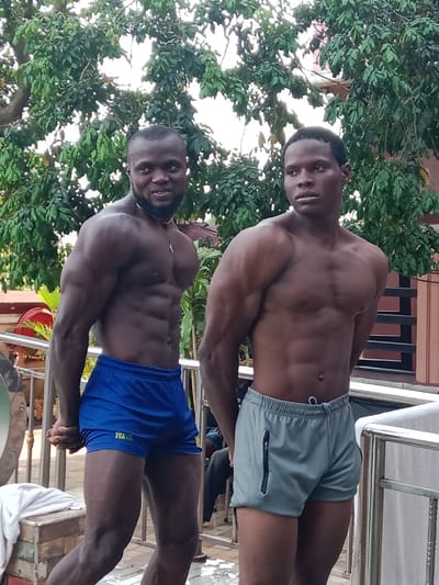 Two muscular men stand side-by-side outside. The man on the left has a trimmed beard and wears blue shorts with "WATA" printed on them. His abs are defined and his arms are muscular. The man on the right wears grey shorts with a zipper on the side. He looks forward with a serious expression. They both have dark skin. Green trees are in the background, along with parts of a building and railing.