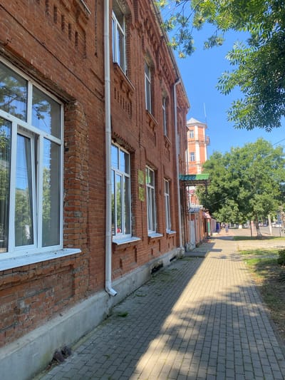 The image depicts a street scene in Maykop, Russia, on a sunny day. A red brick building with white-framed windows lines the left side of the frame. A paved sidewalk runs alongside the building, with shadows cast by trees overhead. In the background, a tall, red and white tower-like structure is visible, along with other buildings and green trees. A few people are walking on the sidewalk in the distance. The overall impression is one of a typical urban street in the summer.