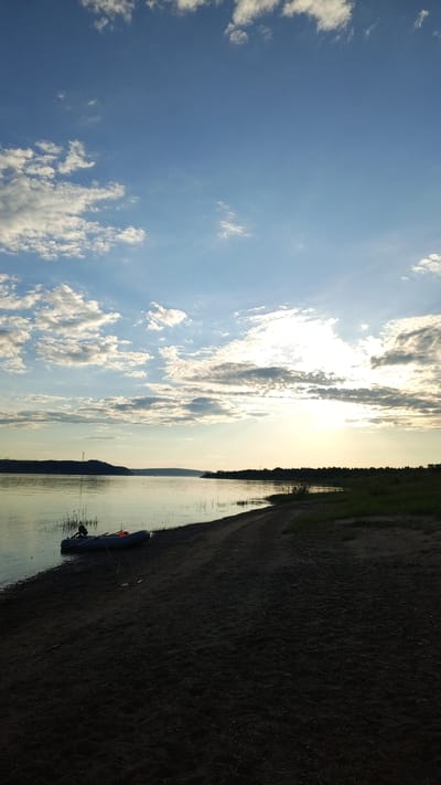The image presents a wide outdoor vista captured during dusk or dawn in Шолья, Russia. The upper two-thirds of the frame is dominated by a clear blue sky interspersed with numerous white stratocumulus clouds, which are particularly dense on the left side and around the sun on the right. The sun, partially obscured by clouds, appears as an intense light source on the right horizon, creating a bright glare and discernible sunbeams. A calm body of water, likely a river or large lake, occupies the mid-ground, reflecting the ambient light and showing minimal surface disturbance. On the distant left bank of the water, a landmass or hill is visible, topped by a distinct, silhouetted structure, possibly a transmission tower. The immediate foreground features a dark, sandy beach curving from the bottom left to the mid-right. A grey inflatable boat with an attached outboard motor rests partially beached on the sand and partially in the shallow water on the left-central side of the foreground. An orange object is visible inside the boat. Thin reeds or tall grass protrude from the water along the near shoreline. The right side of the sandy bank transitions into patches of green grass and low vegetation, leading to a dense line of trees forming the distant right bank. No human figures are discernible in the scene.