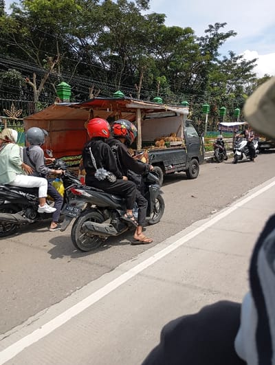 The scene shows a street in Kutawaringin, Indonesia, with a truck parked on the right side of the road, selling fruit. There are several motorcycles parked in front of the truck, with three people on the motorcycles. The person on the far left motorcycle is a woman wearing a white shirt and a green hijab. The person on the second motorcycle from the left is a man wearing a black shirt. The person on the right motorcycle is a man wearing a red helmet. There is a man wearing a white helmet on the truck, facing the back of the truck. The truck is facing forward and has a wooden structure in the back, which appears to be a stall selling fruit. There is a white line separating the right side of the road from the left side, with the truck parked on the right side of the road. The environment is clear and sunny, with a blue sky and some vegetation on both sides of the road. The image is taken from the perspective of a person sitting on another motorcycle, facing forward. The photographer's hand holding the camera can be seen in the bottom left corner.