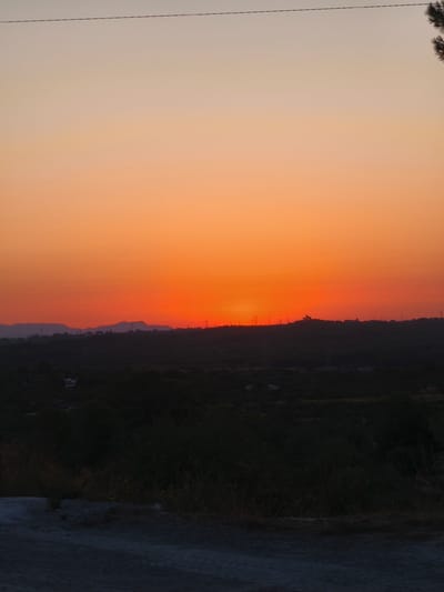 A vertical, eye-level shot captures a sunset over a semi-rural landscape. The sky is a gradient of warm colors, transitioning from pale yellow at the top to a vibrant orange and red near the horizon. A thin, dark power line stretches horizontally across the upper portion of the image. Below the sky, a dark silhouette of rolling hills and dense vegetation forms the mid-ground. Scattered throughout this dark mass are faint outlines of power pylons. The foreground consists of a dirt or gravel road, with dry grass and scrub brush along its edges. The image appears to have been taken from an elevated position, overlooking the town of Riba-roja de Túria, Spain.