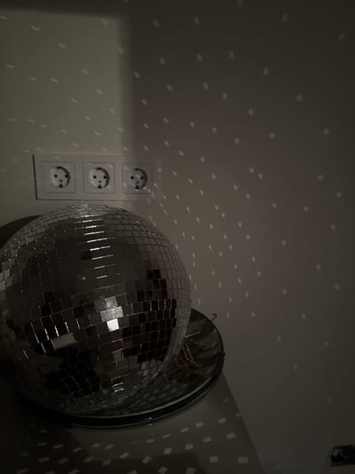 A disco ball sits on a reflective surface on a table in front of a wall with three electrical outlets. The disco ball is reflecting light, casting numerous small, rectangular patterns onto the wall and the surface it rests on. The overall lighting is dim, with the disco ball being the primary source of illumination. The scene appears to be indoors, possibly in a residential or entertainment space. There are no people present in the image.