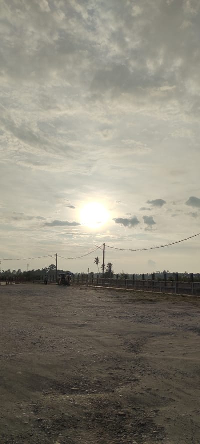 The image depicts an outdoor scene in Meunasahaluedrienlhoksukon Barat, Indonesia, during what appears to be late afternoon or early morning due to the low angle of the sun. The foreground is dominated by a rough, gravelly dirt road. In the middle ground, a fence line runs horizontally across the frame, separating the road from a more distant area. Beyond the fence, several palm trees are visible, suggesting a tropical or subtropical climate. The sky is partially cloudy, with the sun shining brightly through the clouds, creating a diffused light effect. There are utility poles with wires stretching across the scene. On the left side of the frame, near the fence, a person is walking away from the camera, and a three-wheeled vehicle, possibly a tuk-tuk, is present with passengers. The background consists of a treeline and a hazy horizon. The overall impression is a rural or semi-rural setting.