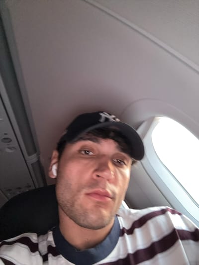 A young man with dark hair is wearing a dark baseball cap and white earbuds. He is looking at the camera and has a striped shirt on. He is in an airplane, as indicated by the airplane window and interior visible in the background. The location is the city of Somma Lombardo, Italy.