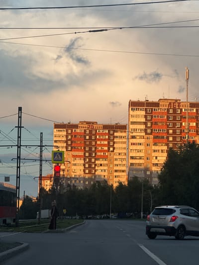 This media file, taken in Magnitogorsk, Russia, captures an urban street scene at dusk. The image is oriented vertically, with the sky and buildings dominating the frame.
The primary subjects are two large, multi-story apartment buildings with a mix of brick and beige facades, featuring numerous windows. A white car is parked on the right side of the road, its brake lights illuminated. In the foreground, there's a complex network of overhead power lines and tram wires, indicative of an urban transportation system. A traffic light shows a red signal, and a pedestrian crossing sign is visible nearby. A person, likely waiting to cross, stands on the sidewalk to the right of the traffic light, partially obscured by shadows.
The setting is a city street with a paved road and a sidewalk. The time of day is late afternoon or early evening, as evidenced by the warm, soft light illuminating the buildings and the darkening sky with scattered clouds. The overall mood is calm and ordinary, depicting a typical urban environment as day transitions into night.
There is some visible text on one of the buildings, appearing to be the number "70" in a large font.
