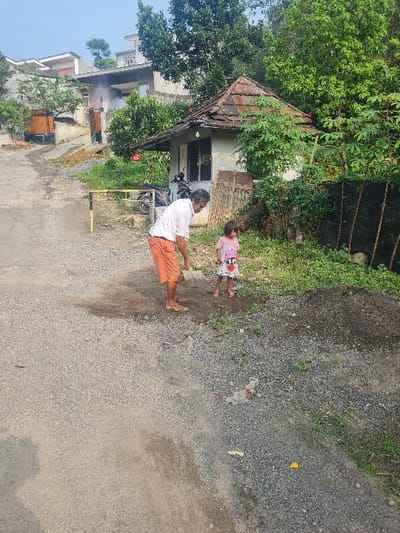 A man in a white shirt and orange shorts stands on a gray road with a child in a pink shirt. The man and child stand near a building with a brown tile roof. Green trees surround the building. A gray wall is to the right. Houses are visible in the background, along with some fencing.