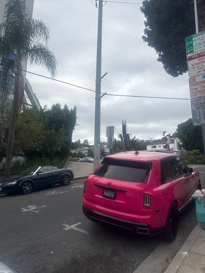 The image depicts a pink Rolls-Royce parked on a street in West Hollywood, United States of America. A black sedan is parked in front of it. There are parking signs nearby. The vehicle's license plate reads 9HWB578. Street markings indicate the direction of parking. The weather appears overcast. A palm tree is visible on the left. In the background, buildings and trees are present. Electrical wires stretch across the sky.