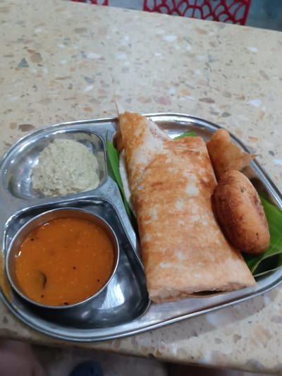 The image displays a close-up, elevated view of a meal served on a stainless steel thali, situated on a speckled countertop surface within a food establishment in Anantapur, India.

The rectangular thali features three distinct compartments. The largest compartment holds two elongated, light brown, folded dosas, positioned partially on a bright green banana leaf section. Adjacent to the dosas is a single deep-fried, golden-brown, oval-shaped item, also resting on the banana leaf. A smaller, circular compartment on the thali contains a light beige, granular chutney. An identical circular compartment holds an orange-red liquid identified as sambar, with several small, dark specks visible on its surface.

The countertop beneath the thali is a light, neutral color, uniformly patterned with embedded irregular fragments of various sizes and muted tones including off-white, grey, and light brown, consistent with a terrazzo finish. In the blurred background, a segment of a red chair back with an openwork, curvilinear design is visible. A dark blue shoe and a partial limb are discernible at the bottom left of the frame, indicating the presence of an individual seated at the table. No direct human interaction with the food is depicted. The scene is static.
