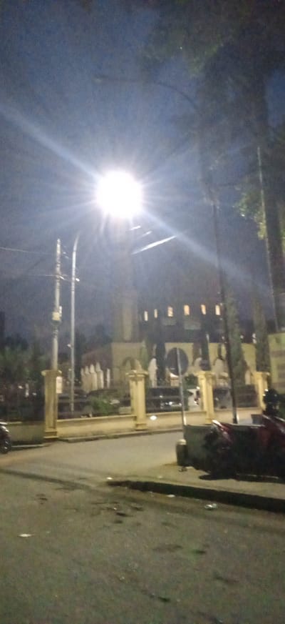 The image captures a nighttime street scene in Banjaran, Indonesia, dominated by a brightly illuminated street light positioned centrally in the upper half of the frame. The light source emits a strong starburst effect, brightening the immediate sky around it against the dark, clear night.
In the background, a large, ornate building stands with multiple small, rectangular, illuminated windows visible along its upper section. Below these windows, the building features distinctive arched designs and what appear to be light-colored figures or statues arranged in front of it, behind a low, decorative fence or wall punctuated by prominent yellow-brown pillars. These architectural elements suggest a public or religious structure, possibly a mosque.
The foreground consists of a dark asphalt road, appearing somewhat uneven or damp, with a visible curb on the right. Part of a red motorcycle or scooter is parked near this curb on the right side. Several utility poles and overhead wires are visible, extending from the left towards the bright street light. Dark foliage from trees can be seen faintly in the upper right corner. The scene is static, with no discernible activity or people in motion. No readable text is visible in the image.