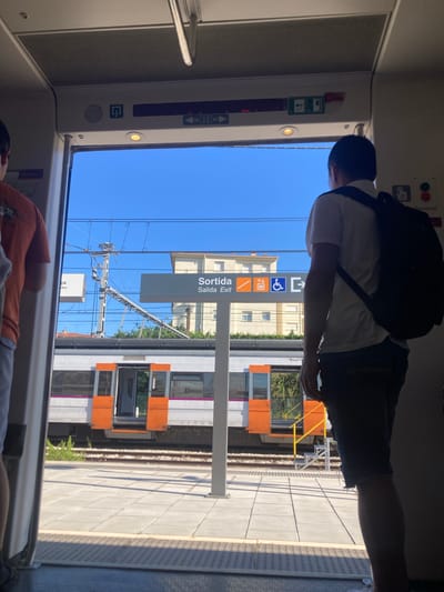 A person wearing a white t-shirt and dark shorts with a black backpack is seen from behind, standing near a train door. Another person in an orange shirt is partially visible, lying down inside the train. The scene appears to be a train station platform, with a train stopped at the platform. The sky is clear and blue, suggesting a sunny day.
Visible text on a sign reads "Sortida" and "Salida Exit," indicating an exit. There are also symbols for disabled access and stairs. The train carriages are white with orange doors. The platform is paved and there are train tracks visible. The overall setting suggests a travel scenario, likely a stop at a station in el Vendrell, Spain. The light suggests it is daytime.