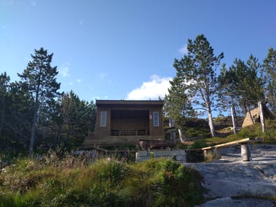 A wooden open-sided shelter is situated on a rise above a rocky, grassy area. The shelter has a wooden floor and benches inside, with windows on either side. A metal grill sits on concrete blocks in front of the shelter, and a wooden bench is positioned on the rocks to the right. Trees surround the shelter, and the sky above is clear and blue. The scene is located in Sveggen, Norway.