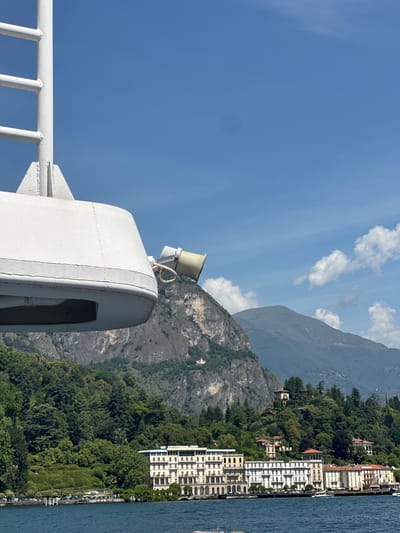 A view from a boat on Lake Como shows a waterfront lined with grand hotels and villas, nestled at the base of lush, green hills. The architecture is classic European, with numerous windows and balconies. The water is a deep blue, and the sky is a clear, bright blue with a few scattered white clouds. In the foreground, the white structure of the boat is visible, including a ladder and a speaker. The scene is tranquil and picturesque, suggesting a summer day in a popular tourist destination. The area depicted is likely the waterfront area of Griante, Italy, known for its beautiful villas and hotels along Lake Como.