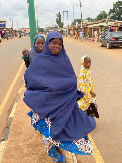 Three females are on a street. Two females are wearing hijabs. One female is wearing a yellow and white top with black pants. The females are on a street in Kafanchan B, Nigeria. There is a black car parked along the street. There are buildings in the background.